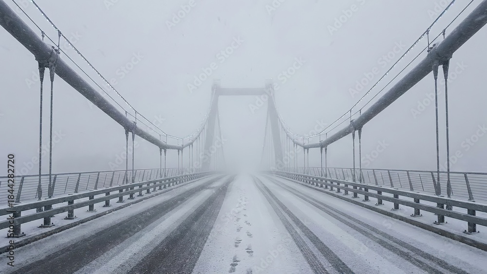 Fototapeta premium Long Suspension Bridge Covered in Fresh Snow and Heavy Fog with Tire Tracks Leading into the Distance, Evoking Winter Travel and Solitude
