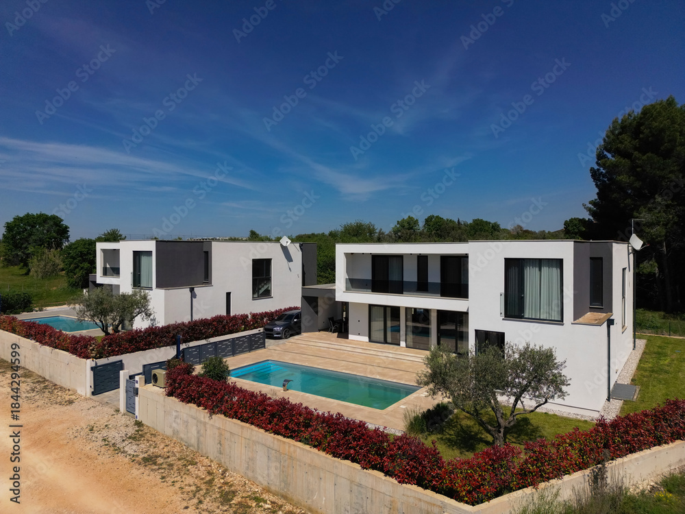 Fototapeta premium Aerial view of two stunning modern luxury villas with private swimming pools, elegant architecture, and lush green landscaping, set against a beautiful natural backdrop