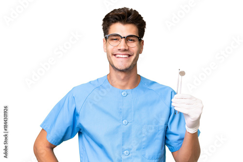 Young dentist man holding tools over isolated background posing with arms at hip and smiling