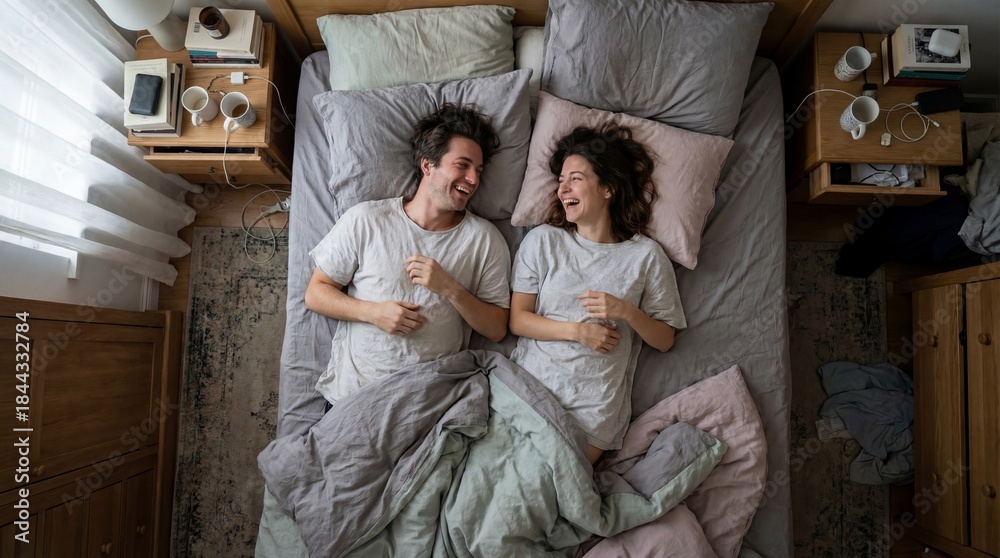 Fototapeta premium Top view of happy couple lying in bed laughing together. Cozy morning scene showing love and relationship intimacy. Ideal for lifestyle content and home comfort concepts.
