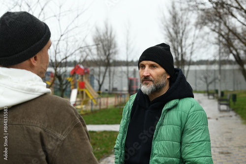 Two men stand facing each other in a park. They are engaged in conversation. The weather is cloudy, and there are playground structures visible in the background