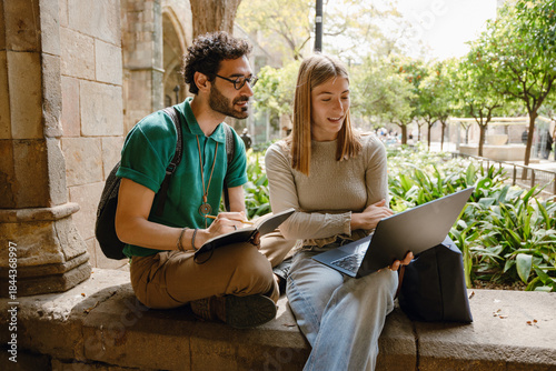 A man and a woman look at a laptop she is showing and sit on a wall while he holds a pencil and a notebook