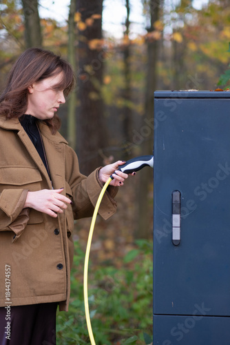 A young woman charges an electric car, trying to navigate the charging station while holding the charging cable. The process of charging an electric car