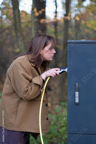 A young woman charges an electric car, trying to navigate the charging station while holding the charging cable. The process of charging an electric car
