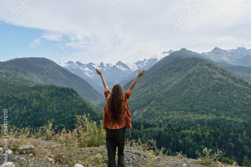 Fotografi A woman stands on a rocky overlook with arms raised, embracing vast mountains and green valleys in a dramatic nature landscape