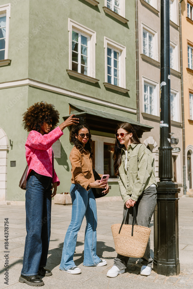 Fototapeta premium A woman is taking a picture on her phone while two female friends are standing next to her, looking at the phone that one of them is showing while one of them is leaning on a street lamp