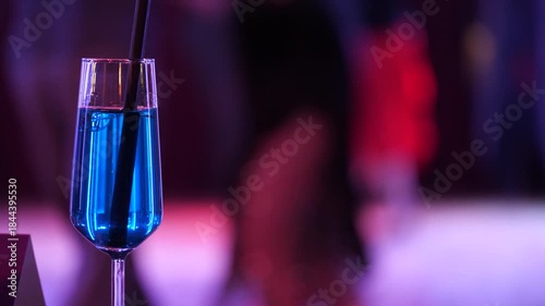 A glass of blue cocktail takes center stage. A glass of cocktail against a blurred ballroom dance floor. Couples dancing in the background