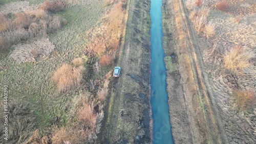 Aerial view of a car riding an off-road path alongside a river at late autumn. SUV driving along rural route. Auto navigating surrounded by dense foliage and uneven terrain. Adventure and ruggedness