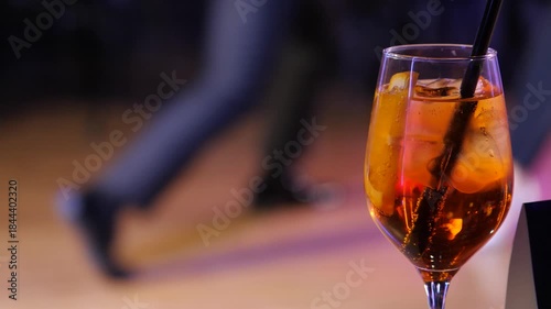 A cocktail takes center stage. A crystal glass with citrus and ice against a blurred ballroom dance floor. The feet of a dancing couple are in the background. Slow motion 2x