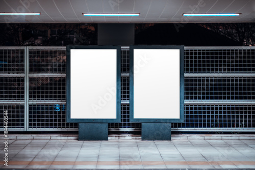 Urban mock-up scene with two blank vertical billboard posters templates at an outdoor subway platform, illuminated frames against metal grid wall, tiled floor and fluorescent lights