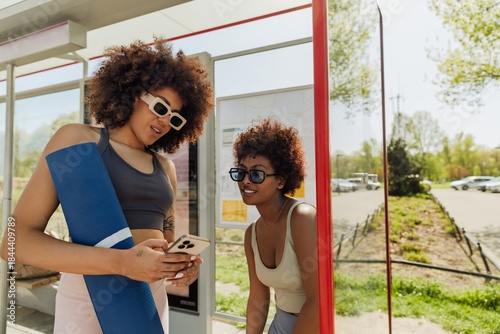 Woman holding a mat and talking while showing her phone to her female friend who is smiling and leaning on a bus stop