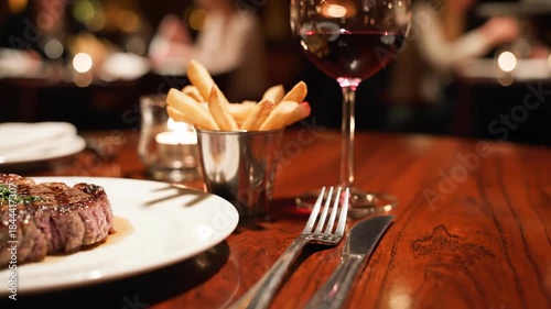 Elegant slow pan across a polished dark wooden restaurant table setting, highlighting the perfectly grilled steak and fries in shallow depth of field ambient lighting Elegant, dining, ambient
