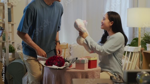 Romantic happy couple in love exchanging valentine gifts at home, smiling faces showing affection on special day with beautiful roses, presenting fluffy bear to partner.
