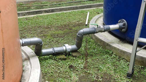 A gray PVC pipe connection, including a green ball valve, linking two large outdoor plastic water storage tanks orange and blue on a grassy area with concrete curbs.