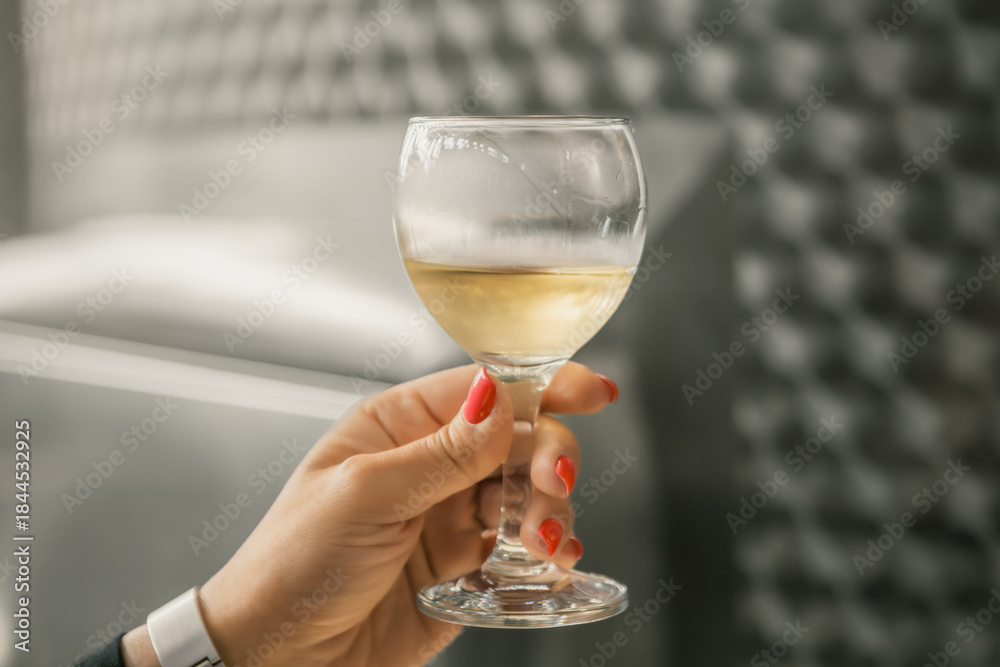 Obraz premium A close-up of a hand holding a glass of white wine in a winery setting, with stainless steel fermentation tanks and industrial lighting in the background, creating a refined and elegant atmosphere.