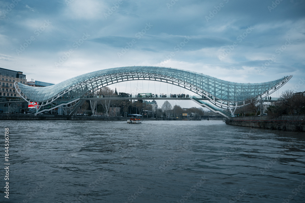 Obraz premium The Bridge of Peace in Tbilisi, Georgia, features a modern bow-shaped glass and steel canopy spanning over the Kura River under a cloudy sky