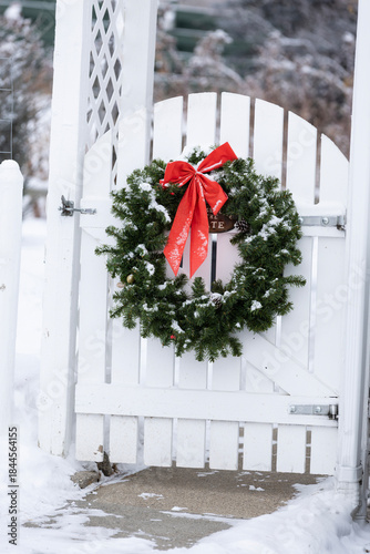 A festive green Christmas wreath with a red bow hanging on a white wooden garden gate in the winter.