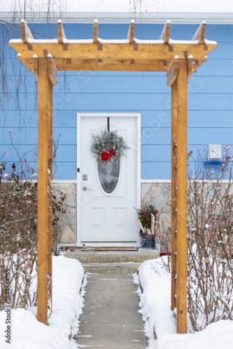 Festive green Christmas wreath decoration hanging on a white door of a blue house with a wooden arch in the winter.