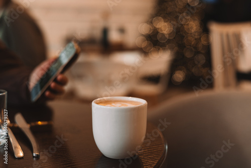 A cozy, warm scene of a person enjoying a cup of cappuccino in a modern cafe during the winter holiday season. The blurred Christmas tree in the background adds a festive touch to this casual scene.