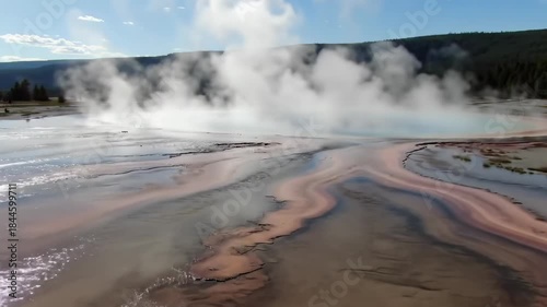 Colorful geyser with steam rising, trees in the background, clear blue sky with clouds on a sunny day