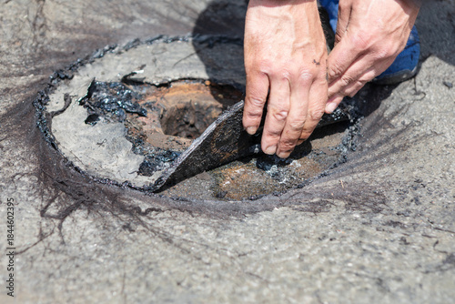 Worker lifts deteriorated roofing material during flat roof drainage system repair, exposing rusted metal and tar-coated layers. Closeup highlights inspection and maintenance of damaged roof component