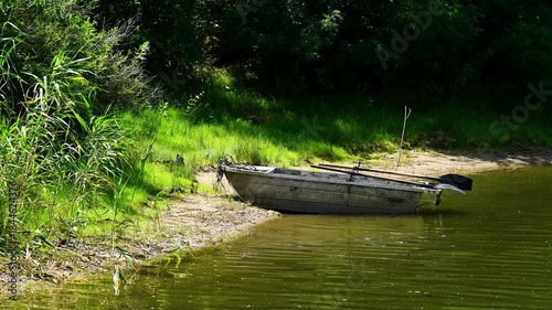 An old wooden boat tied to the shore of a small lake or river in a rural or wooded area. Summer natural landscape.