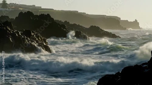 Crashing waves hitting rocky coast during golden hour create misty sea spray and a silhouetted headland backdrop