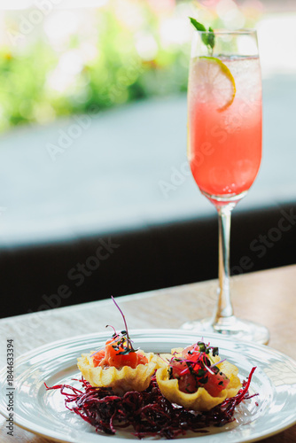 Elegant appetizer plate with tartlets and vibrant cocktail garnished with lemon and mint on a wooden table in a bright setting