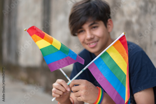 Teenaged boy wears rainbow wristband and holding rainbow flags, lgbtqai symbol, sitting in front of wall, concept for calling out all people to respect human rights of lgbtqai people around the world.