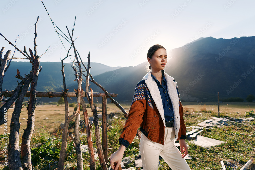 Fototapeta premium Woman in sweater and jacket standing by a rustic fence in countryside mountains at golden sunlight. Outdoor portrait, travel lifestyle with contemplative pose and natural rural scenery.