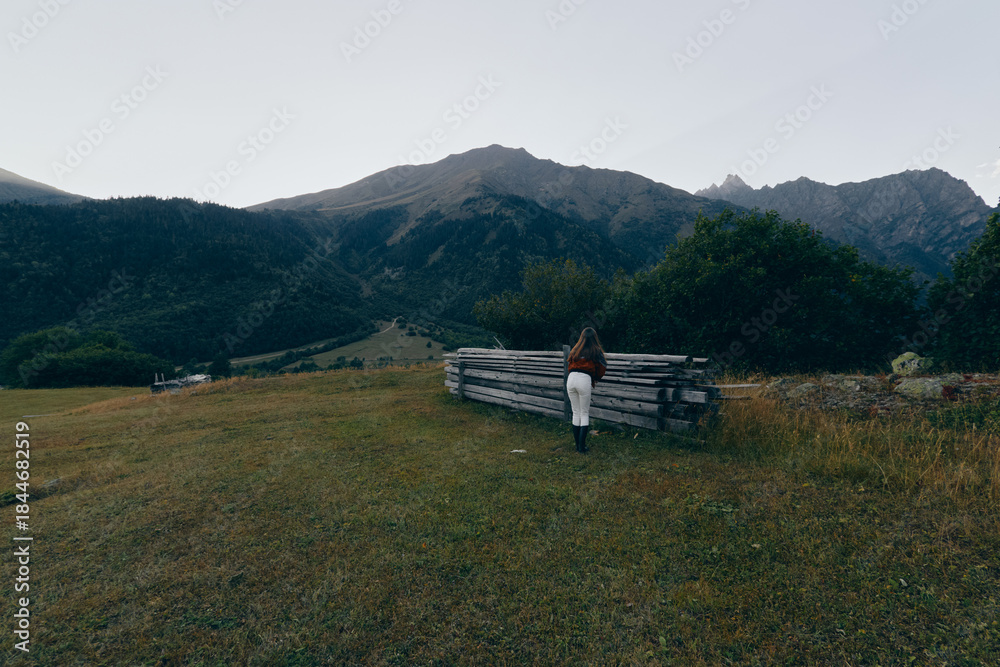 Fototapeta premium Mountains meadow fence person backpack hiking landscape solitude grass nature. Lone hiker walks beside a wooden fence across a grassy field toward forested mountain peaks under moody sky.