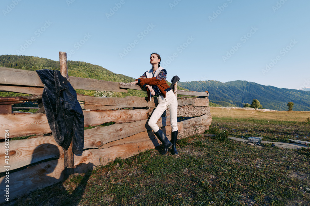 Fototapeta premium Woman leaning on wooden fence in countryside mountains holding a hat and jacket, wearing boots and casual outfit outdoors, enjoying scenic meadow, freedom and relaxed travel mood.
