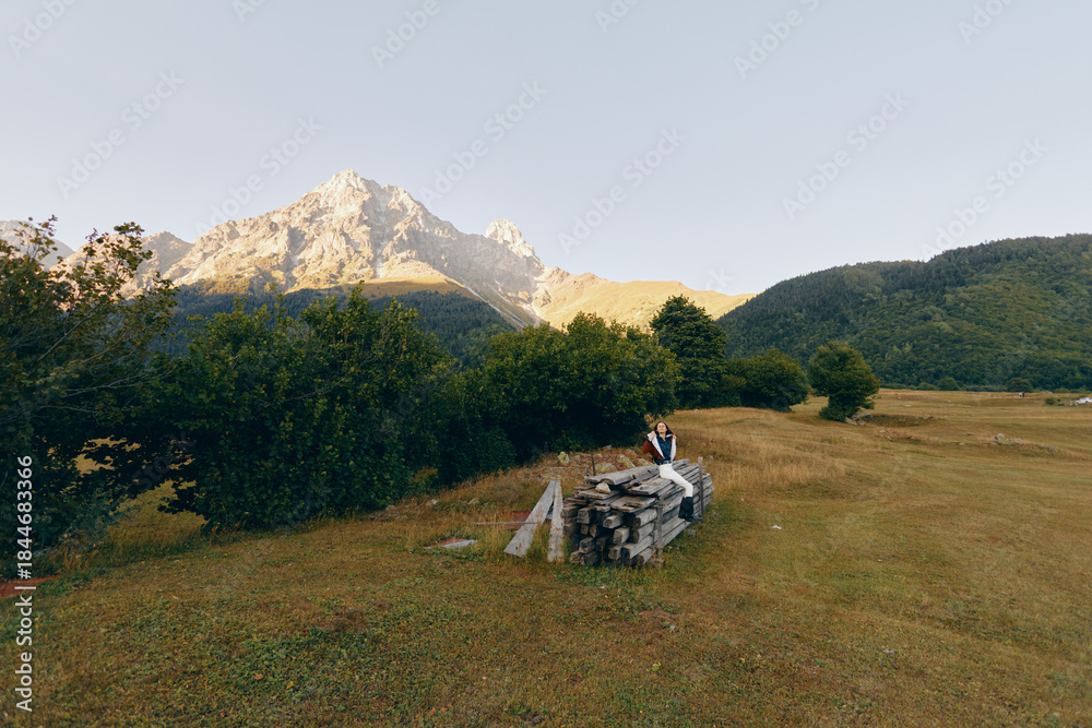 Fototapeta premium Mountain meadow with hiker and backpack sitting on a stonewall, trees and distant peaks at sunrise. Landscape scene of nature, solitude, outdoor travel and hiking adventure.