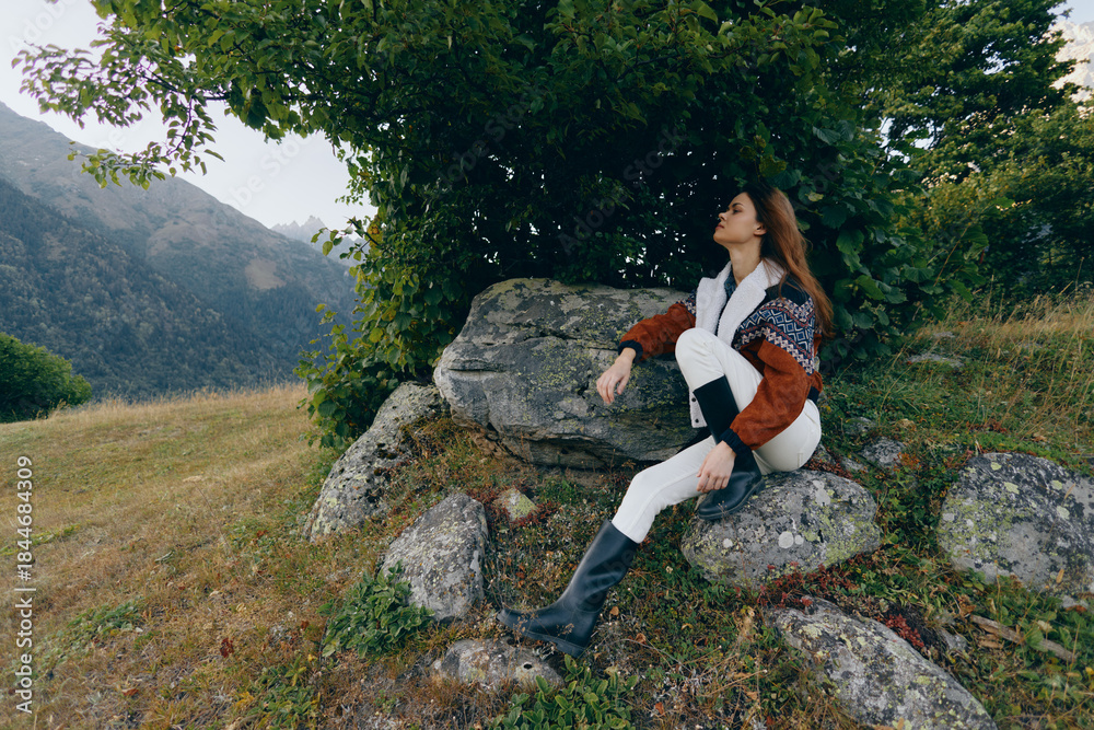 Fototapeta premium Woman sitting on rocks under a tree in a mountain meadow with mountains in the background, nature scenery and serene landscape, relaxed portrait in sweater and boots enjoying peaceful relaxation.