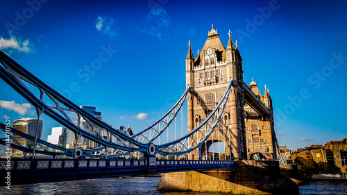 Tower Bridge captured from a low-angle perspective under clear blue skies, highlighting its iconic architecture and the modern London skyline in warm afternoon light.

