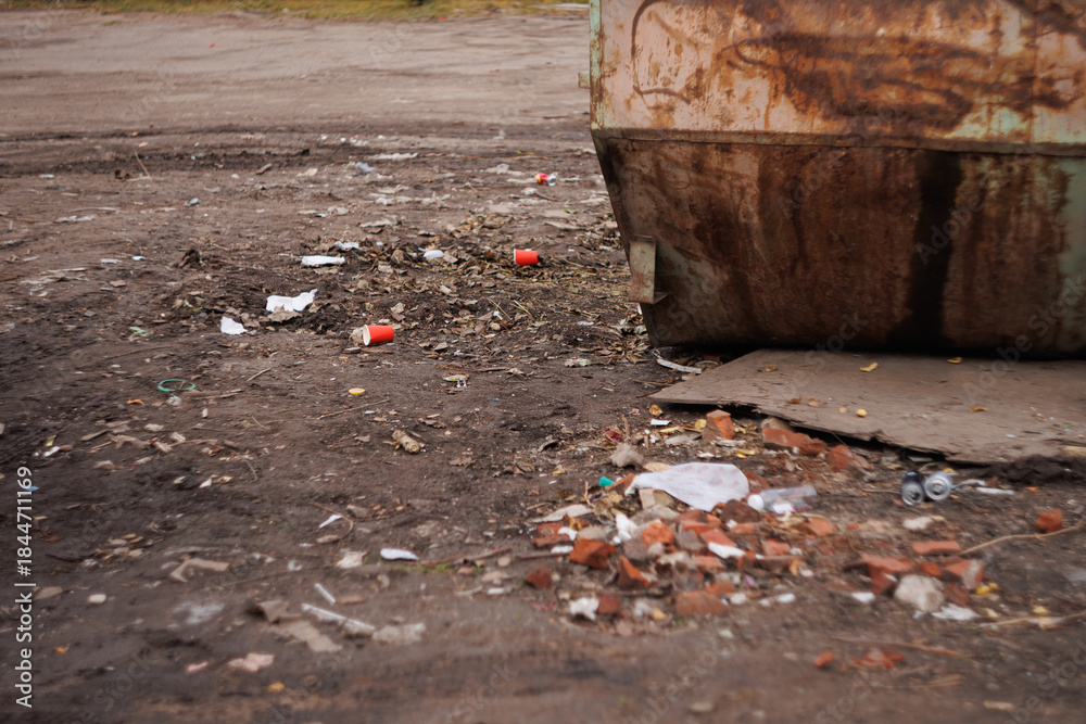 Obraz premium Litter scattered on dirt ground near rusty metal dumpster outdoors. Urban waste scene with cups and debris. Pollution, neglect, environment, cleanup concepts.