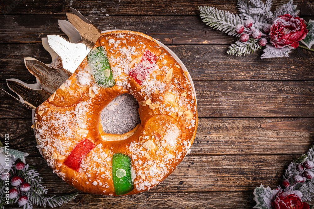 Naklejka premium Roscon de reyes with cream and christmas ornaments on a wooden background, top view. Roscon is a traditional Spanish Christmas pastry cake with decoration. Kings day concept spanish three kings cake.