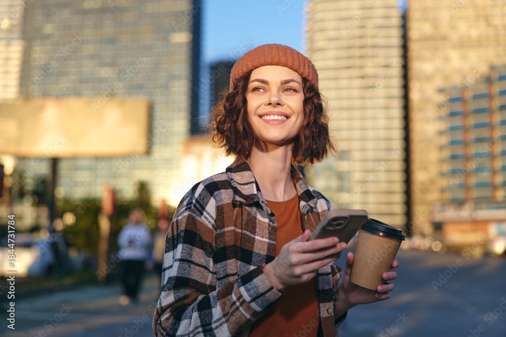 Obraz premium woman coffee phone smile urban beanie sunlight candid Young woman in a beanie holds coffee and phone on a city street, smiling in golden hour glow; authenticity, mindful living and emotional