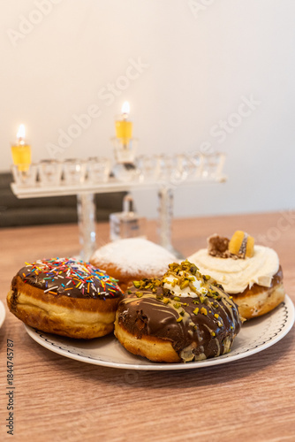 A festive plate of Hanukkah sufganiyot (filled doughnuts) with assorted toppings sits on a wooden table, lit by the first candle on a menorah in the soft background.