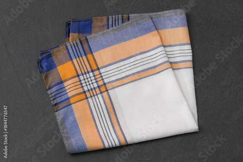 A neatly folded white handkerchief with a yellow, blue, and black plaid border on a dark gray, textured surface.