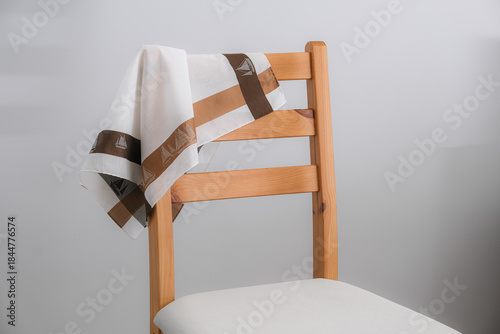 A white cotton handkerchief with a wide brown and tan border, featuring a sailboat pattern, is casually draped over the back of a light wooden chair with a white padded seat.