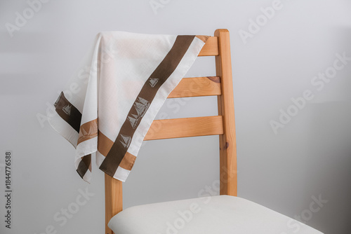 A white cotton handkerchief with a wide brown and tan border, featuring a sailboat pattern, is casually draped over the back of a light wooden chair with a white padded seat.