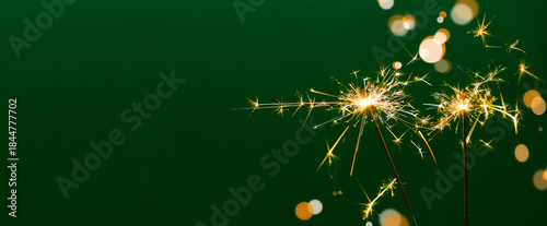 Bright golden sparks from two burning sparklers against a deep emerald green background with soft bokeh. A perfect background image for Christmas cards, New Year's banners, and holiday designs.