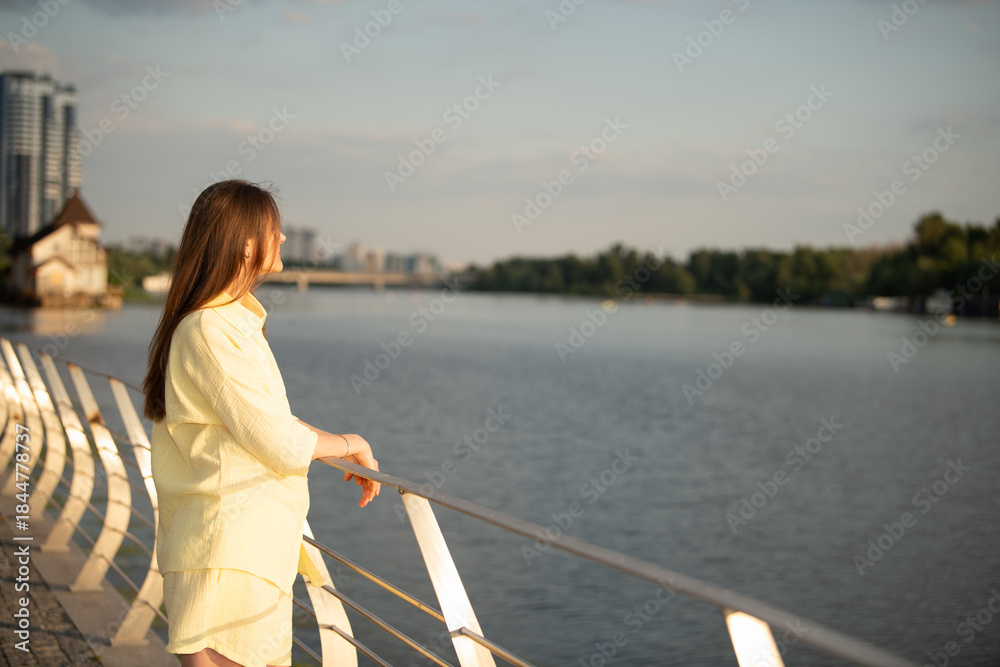 Fototapeta premium A woman stands by a white railing with a river in front. She looks out at the water and city buildings in the background. The scene is in the late afternoon