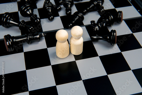 Two figures standing together on chessboard among fallen pieces representing partnership, unity, trust, and strategic alliance facing challenges in competitive environment.
