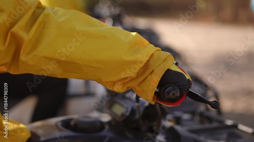 Gloved hand on handlebar throttle preparing lineup of quads, yellow jacket sleeve and textured grip in sunlight, twisting motion signals imminent start, shallow