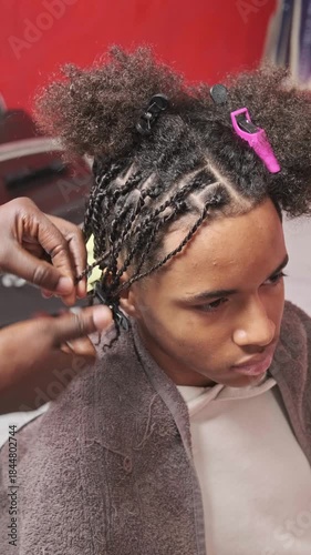 Smiling teen boy from slight top-front view as stylist perfects braids on the right side of his afro hair