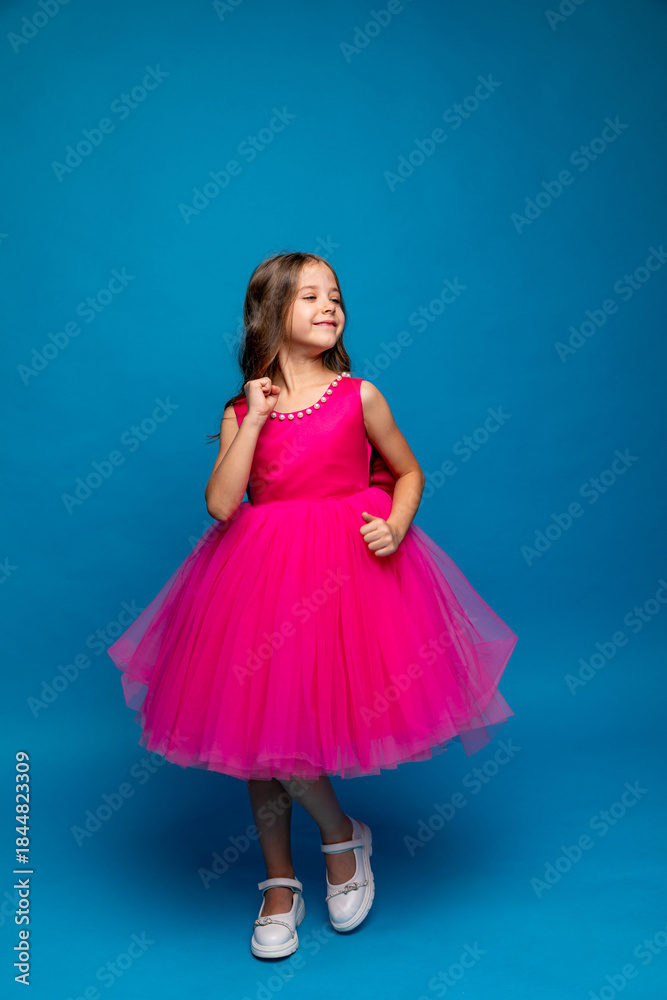 Naklejka premium Cute happy little girl in a pink fluffy princess dress has fun and dances on blue background. Kids Birthday party celebration concept. Happy Birthday, copy space. High quality photo