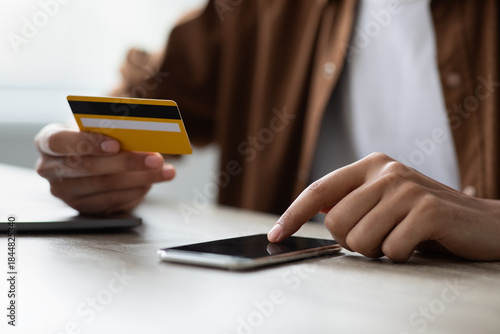 Valokuva A person holds a yellow credit card in one hand while using their finger to interact with a smartphone on a wooden desk
