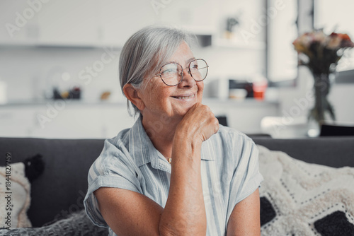 Elderly woman sitting on a sofa at home smiles as she looks out the window. She enjoys a peaceful and reflective moment, embracing a calm and cozy domestic lifestyle.